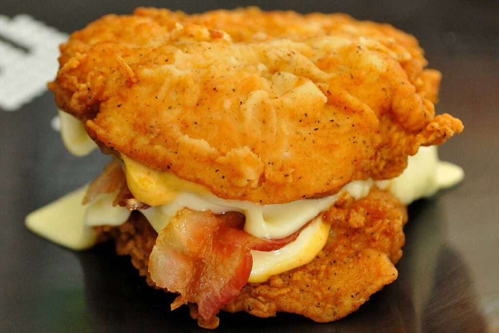 A sandwich featuring crispy fried chicken fillets as buns, filled with bacon, melted cheese, and sauce. This innovative creation, reminiscent of discontinued fast food items, is served on a dark, reflective surface.