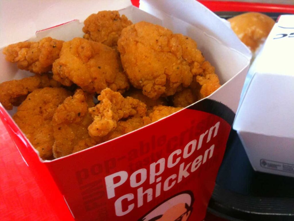 A red and white box labeled Popcorn Chicken, filled with crispy, golden-brown pieces—a nostalgic treat reminiscent of discontinued fast food items. The box rests on a red tray with the edge of another food box peeking from the background.