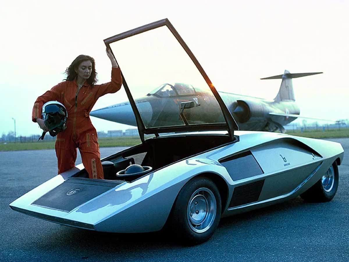 A Lancia Stratos Zero (1970) concept car on the tarmac with a woman stepping into the car and a fighter jet behind her