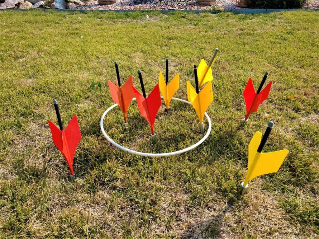 A set of red and yellow lawn darts is spread out on the grass, with many landing inside a white circular target. The scene is outdoors on a sunny day, with gravel and plants visible in the background.