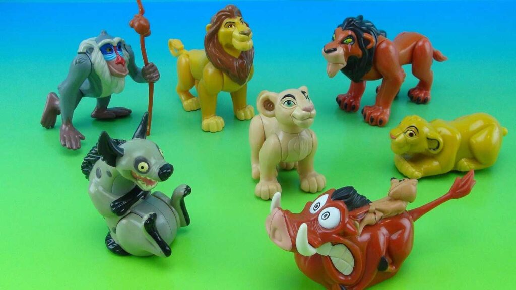 Toy figures of animated animal characters, including a lion, meerkat, warthog, hyena, baboon, and lion cub, arranged on a green and blue gradient background.