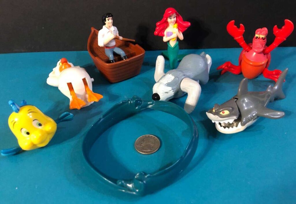 A collection of plastic toys depicting a mermaid, a man in a boat, a lobster, two fish, a bird, a dog, and a shark are arranged around a coin on a blue surface.