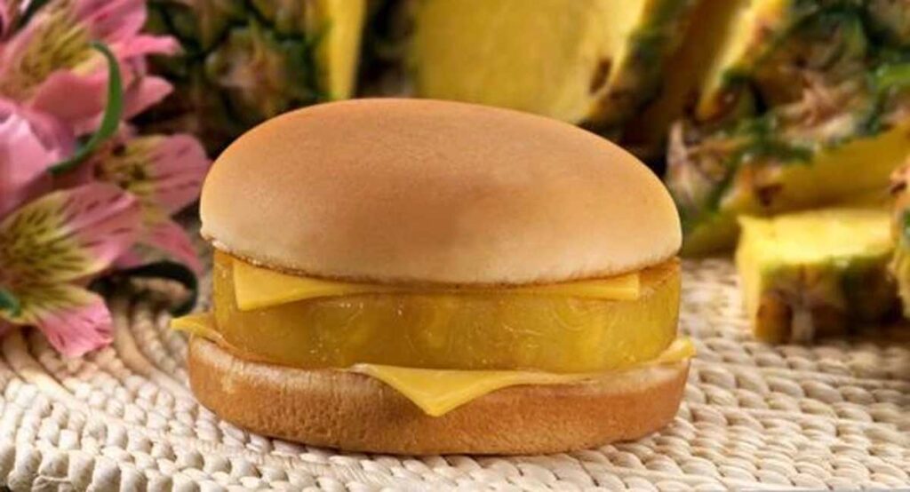 A sandwich featuring a pineapple slice and cheese nestled in a bun is set against a background of pineapple pieces and pink flowers, reminiscent of beloved discontinued fast food items.