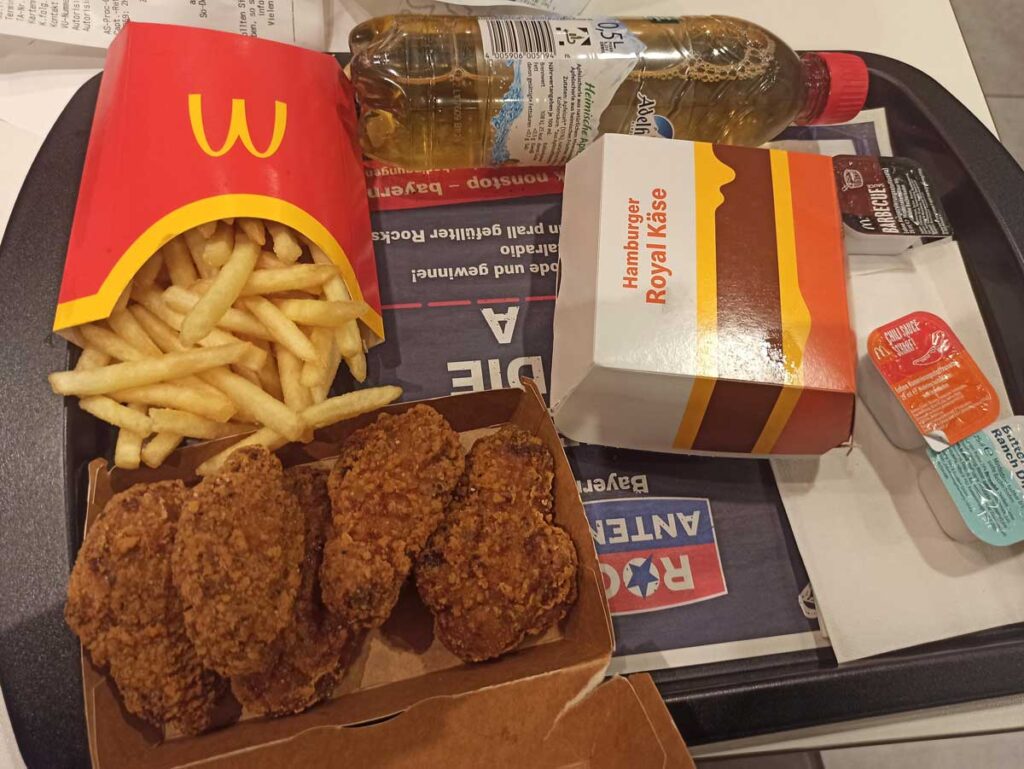 A fast food tray with a McDonald's meal: a box of chicken nuggets, a hamburger, fries in a red container, and apple juice. Among the sauces, reminiscent of discontinued fast food items, are two dipping packets—one red and one light blue.