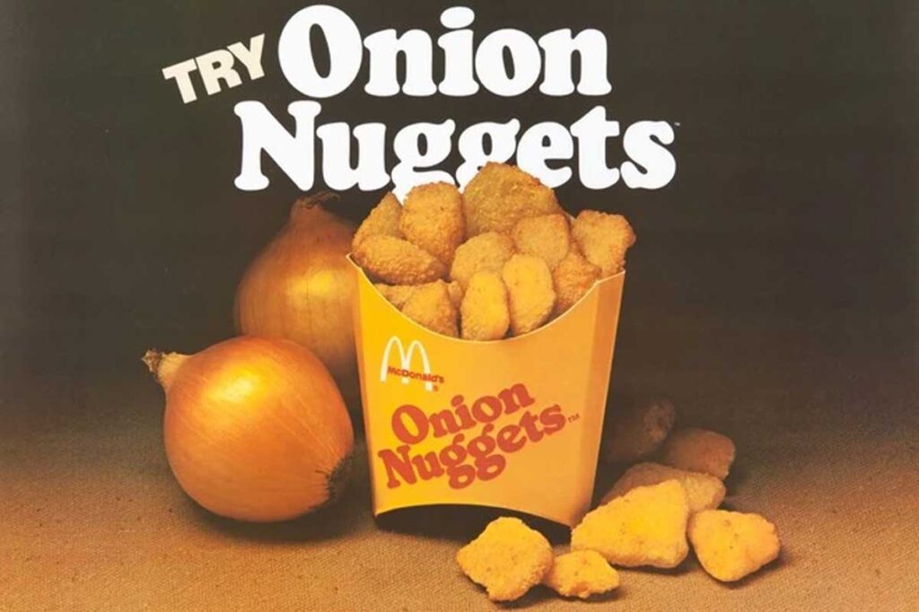 A vintage McDonald's advertisement showcases the now-discontinued fast food item, Onion Nuggets, elegantly served in a branded container and surrounded by whole onions. The text reads, "Try Onion Nuggets" in bold white letters against a dark backdrop to emphasize the product.