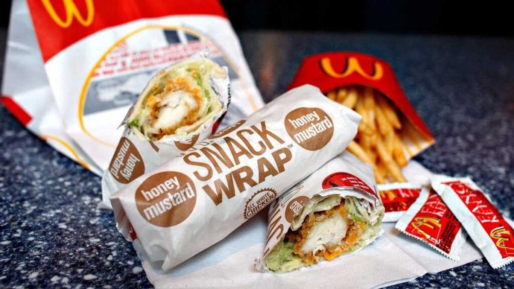 A wrap with chicken and lettuce is partially unwrapped on a table next to a packet of fries and ketchup sachets, reminiscent of some discontinued fast food items. McDonald's branding is visible on the packaging.