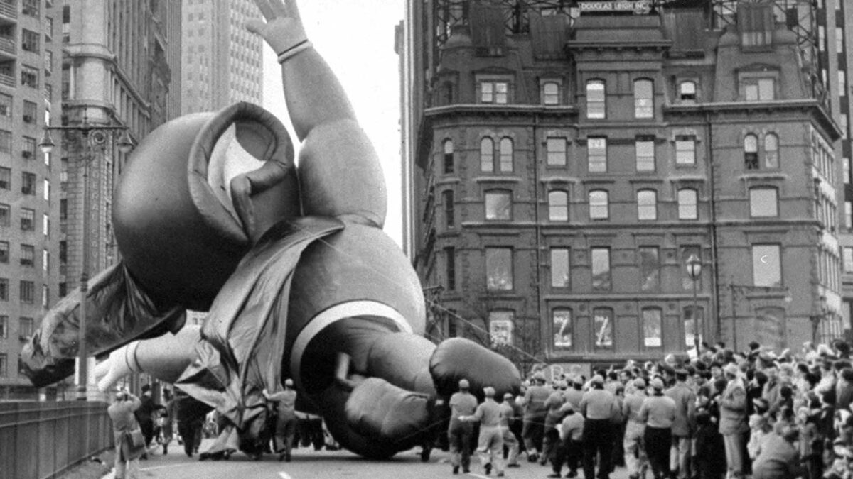 A large balloon figure deflates and falls over a crowded city street during a parade, surrounded by buildings and onlookers. The scene captures a moment of surprise and chaos as people gather around the scene.