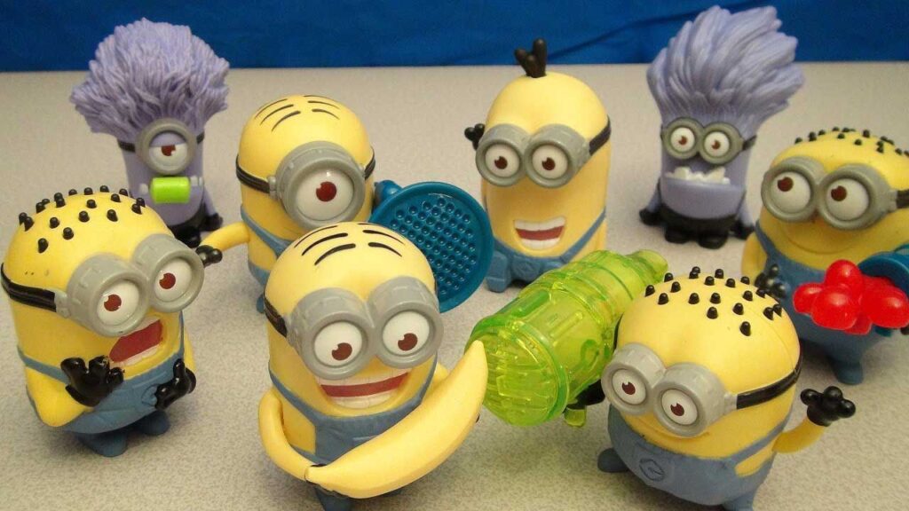 Seven Minion toys, each with unique expressions and accessories like bananas and flowers, are gathered together. Two have purple hair, and all are wearing their signature goggles and blue overalls. They appear playful and lively.