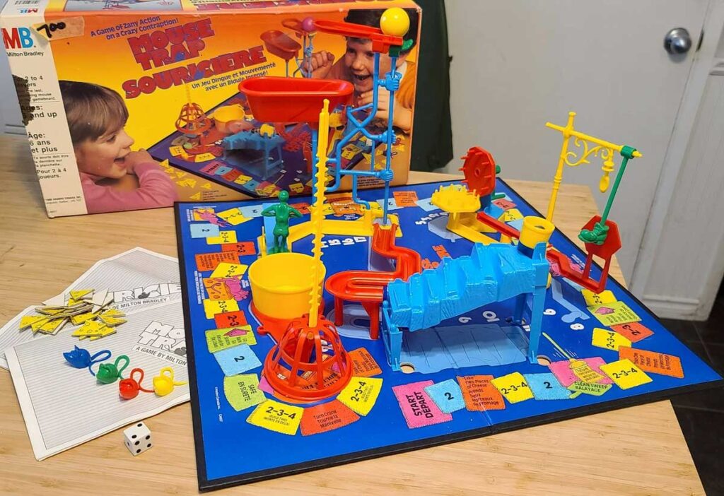 A colorful board game setup of Mouse Trap, featuring a complex contraption with gears, ramps, and a basket. The board is surrounded by a die, yellow tokens, and an instruction booklet. The game box is in the background.