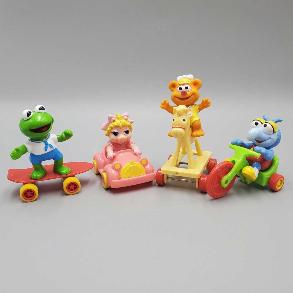 Four colorful toy figures are shown: a frog on a skateboard, a pig in a pink car, a bear on a yellow rocking horse, and a blue character on a red tricycle. They are set against a plain gray background.