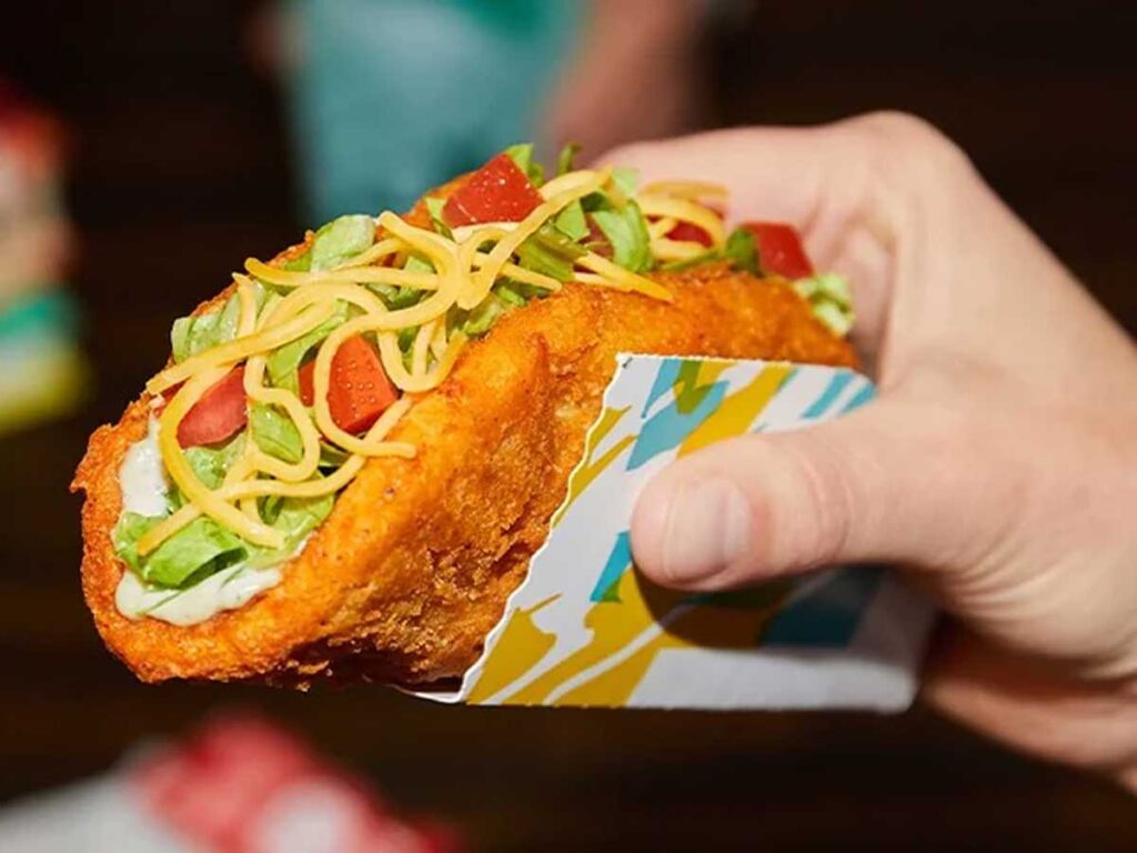 Close-up of a person holding a discontinued Taco Bell menu item: a taco with a crispy shell. It's filled with shredded cheese, lettuce, diced tomatoes, and creamy sauce, all wrapped in colorful packaging.