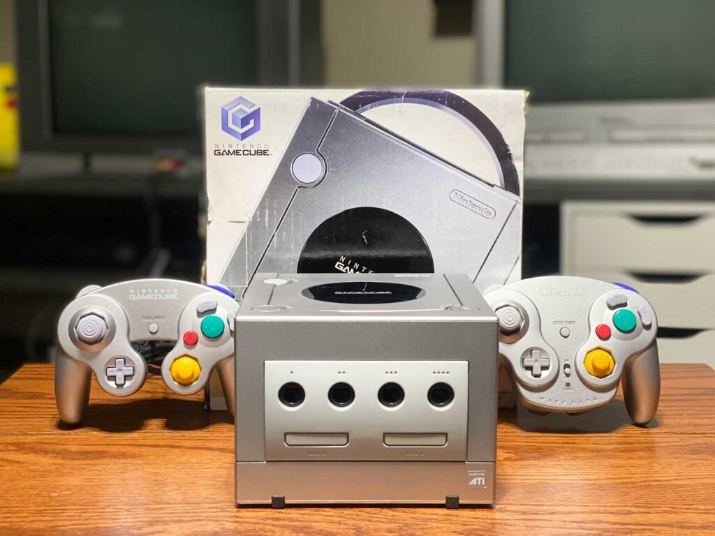 A silver Nintendo GameCube console, one of the best-selling video game consoles, is displayed on a wooden surface with two matching controllers. The original box with the iconic GameCube logo proudly stands behind the console.