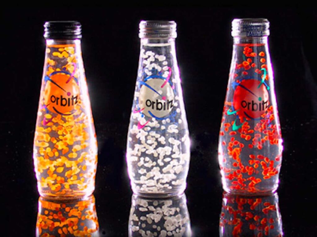 Three glass bottles of the iconic Orbitz soft drink, a beloved discontinued snack from the 90s, are displayed against a black background. Each bottle contains brightly colored floating spheres: orange, clear, and red. The transparent liquid creates a visually striking effect with the suspended spheres.