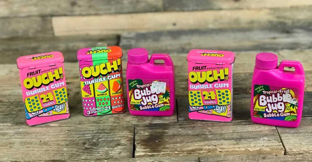 Assorted pink packaging of nostalgic 90s bubble gum products, including discontinued snacks from the 90s like Ouch! and Bubble Jug, featuring colorful fruit-themed designs, displayed against a wooden backdrop.