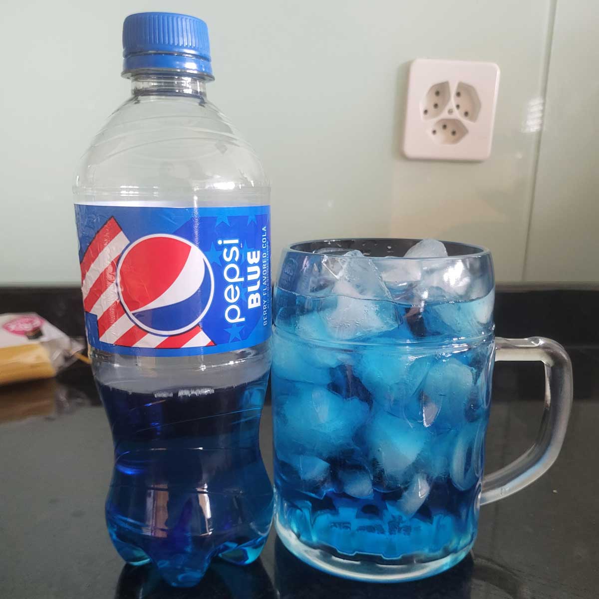 A half-empty plastic bottle of Pepsi blue sitting next to a glass mug of Pepsi Blue over ice.