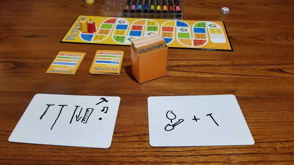 A Pictionary board game setup on a wooden table. There are cards, a die, markers, and a board. Two dry-erase boards show drawings: one with lines, a hammer, and ruler; the other with a circle, line, and the letter T.