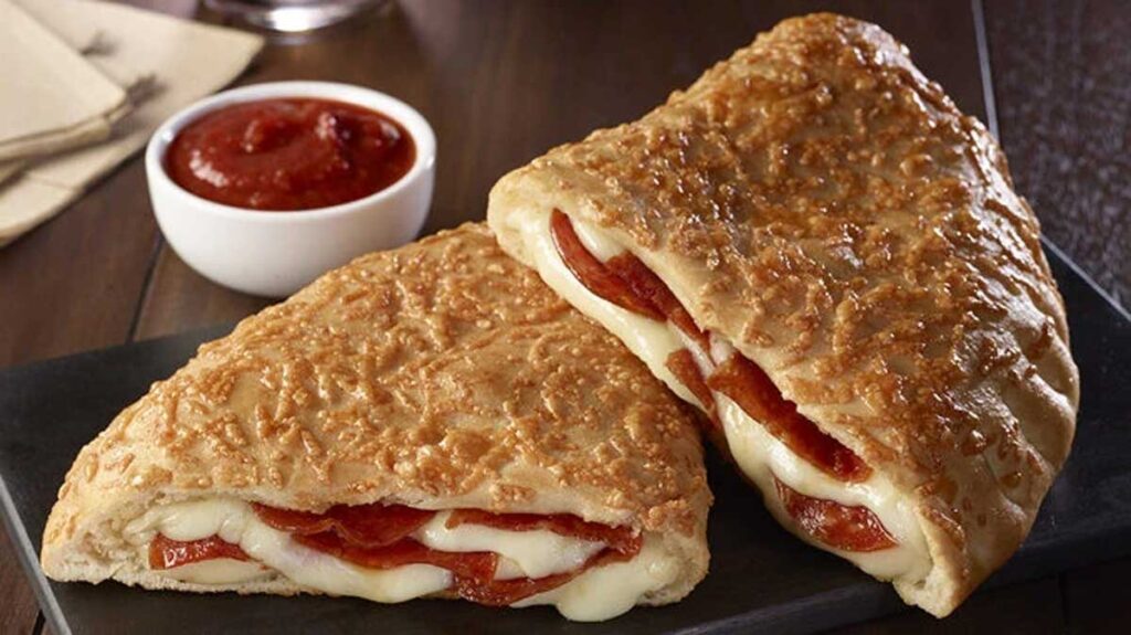 Two calzones filled with melted cheese and pepperoni are placed on a dark surface, reminiscent of beloved but discontinued fast food items. A small bowl of marinara sauce is on the side, set on a wooden table.