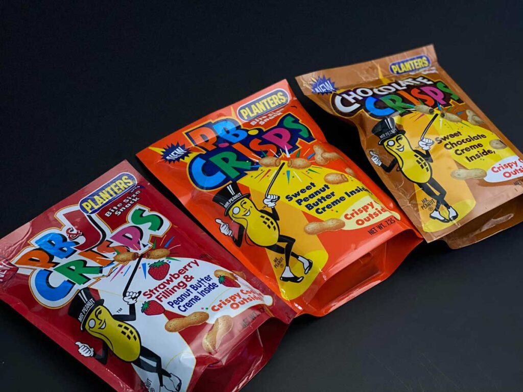 Three colorful snack pouches labeled PB CRISPS by Planters, iconic discontinued snacks from the 90s, are lined up. Flavors include Strawberry, Peanut Butter, and Chocolate, each with a Mr. Peanut character holding a crisp. Packages are displayed on a dark surface.