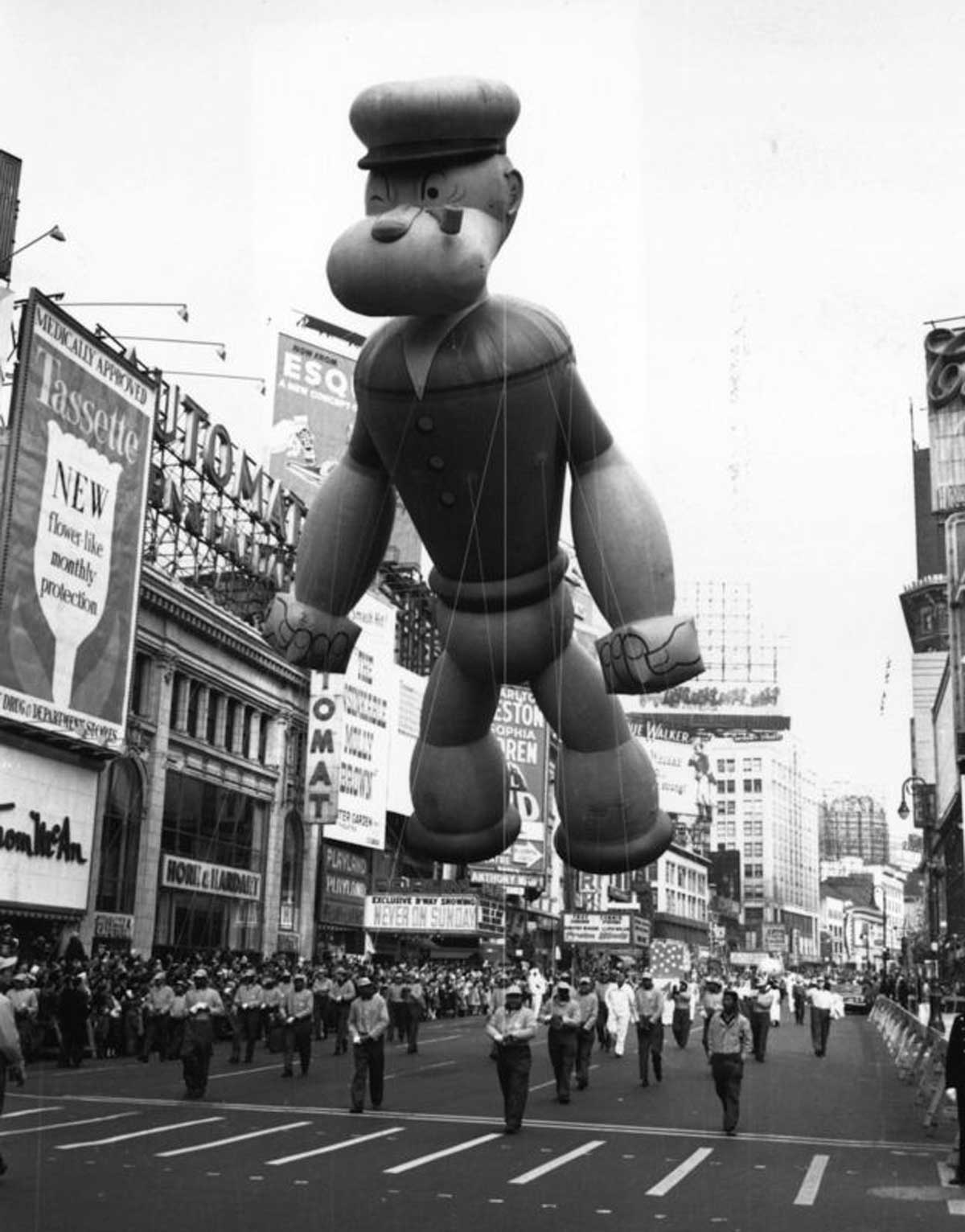 A giant cartoon character balloon floats above a busy street during a parade. People walk below, and buildings with large advertisements line the street. The scene captures a lively, festive atmosphere.