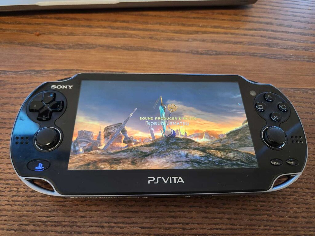 A best-selling PlayStation Vita handheld console rests on a wooden surface, showcasing a fantasy-themed video game screen with rocky landscapes and a sword embedded in the ground.