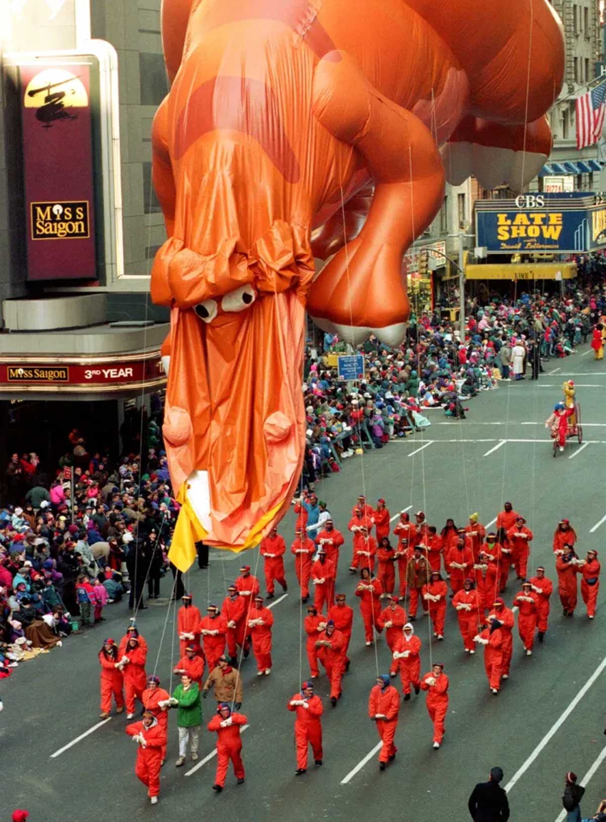 Parade scene featuring a large orange dinosaur balloon and many people in red outfits guiding it. Spectators sit along the street near theater signs.