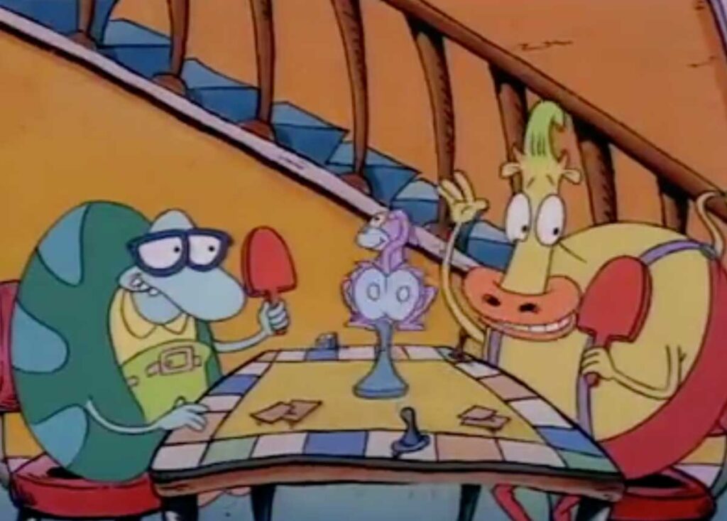 Two cartoon characters, a turtle and a cow, sit at a table in a colorful room. They hold table tennis paddles and appear excited. The table is set with a centerpiece and a tablecloth with a checkered pattern.