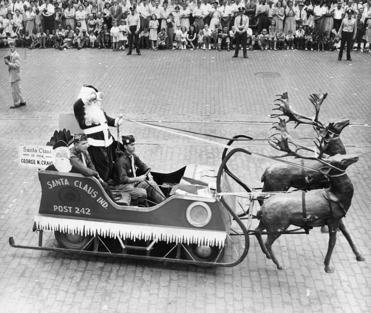 A black and white photo of a parade featuring a Santa Claus riding in a sleigh pulled by reindeer. The sleigh is labeled Santa Claus Ind. Post 242. Crowds of people stand on the sidewalk watching the procession.