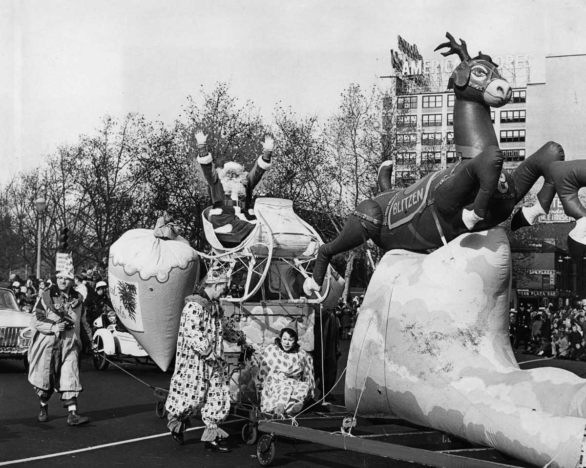 Gimbel's Thanksgiving Day Parade - A black and white photo of a festive parade featuring a large inflatable reindeer pulling a sleigh with a Santa Claus figure. Clowns walk alongside, and a crowd watches from the background. Buildings and trees line the street.