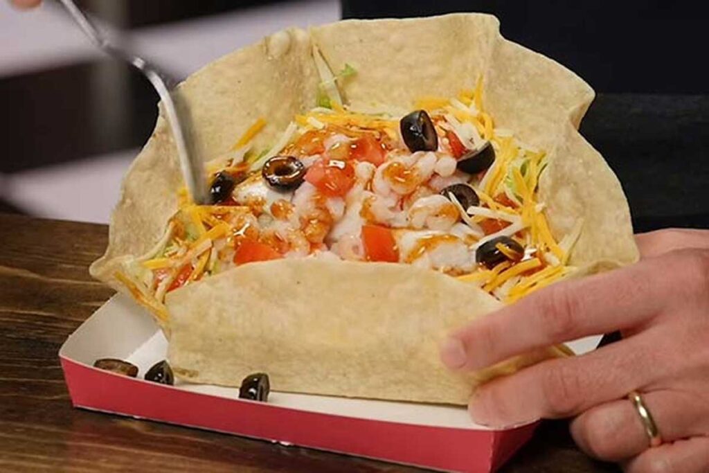 A person is holding a taco salad reminiscent of discontinued Taco Bell menu items in a tortilla bowl on a red tray. The salad is topped with shredded cheese, diced tomatoes, olives, and a drizzle of sauce, with a fork to the side.