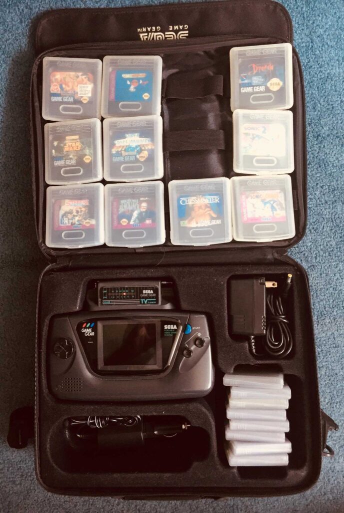 An open Sega Game Gear carrying case showcases one of the best-selling video game consoles. Inside, a Game Gear console is flanked by multiple game cartridges in plastic cases, an AC adapter, and several empty game cases, all meticulously arranged on a blue surface.