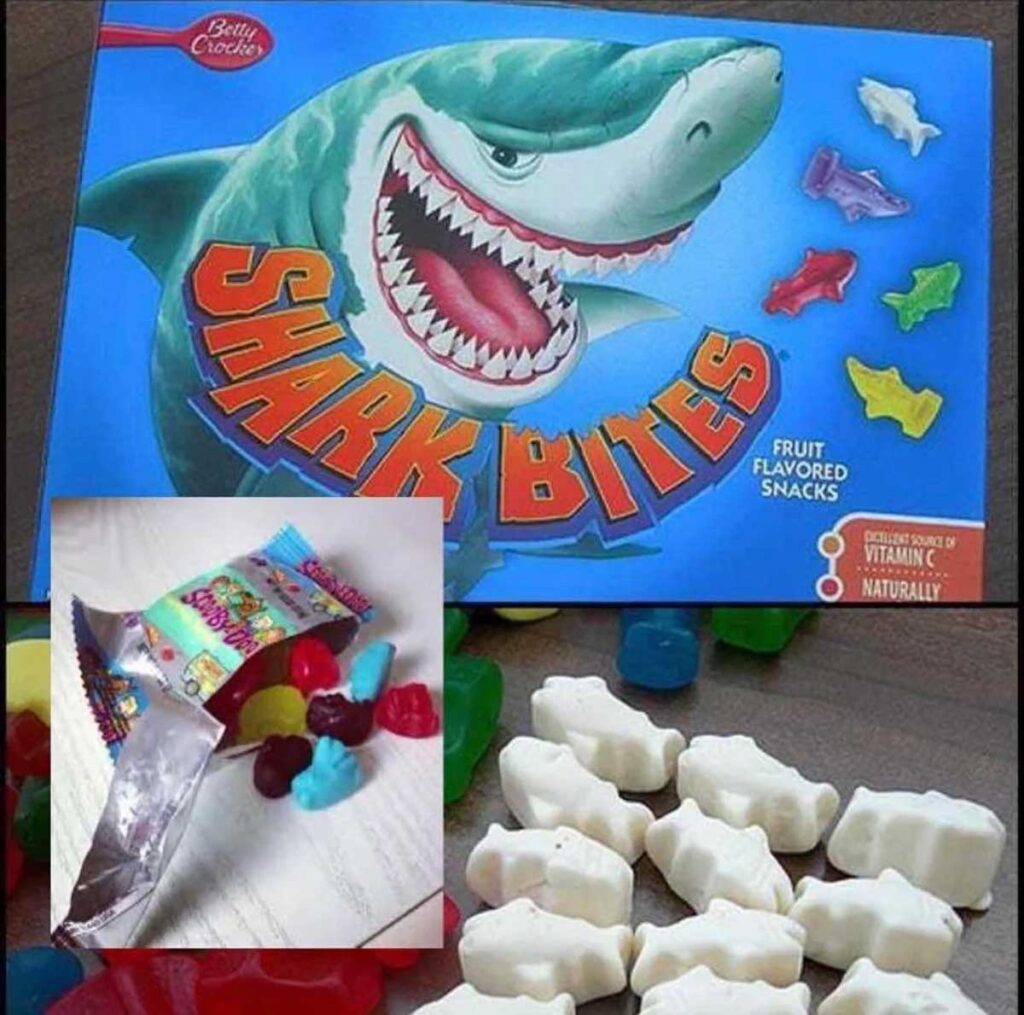 A box of Shark Bites fruit-flavored snacks featuring a large shark graphic harks back to discontinued snacks from the 90s. An inset image reveals open packaging with vibrant gummy candies and several white, shark-shaped gummies.