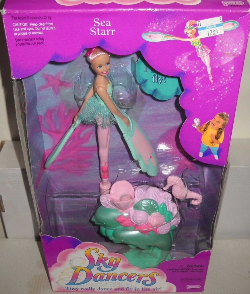 A Sky Dancers doll named Sea Starr is displayed in its packaging. The box is pink and purple with illustrations of the doll flying. The doll wears a green outfit with pink accents and stands on a green base. Price tag visible.