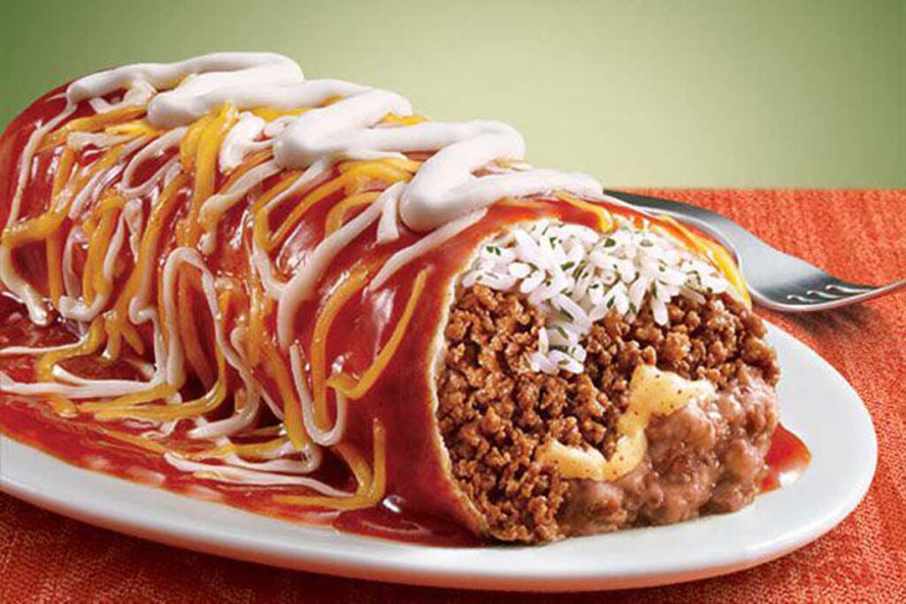 A close-up of a large burrito cut open reveals ground beef, rice, and beans inside. Topped with melted cheese, red sauce, and sour cream drizzles like those beloved but discontinued Taco Bell menu items, it's served on a white plate with a fork by the side against a green background.