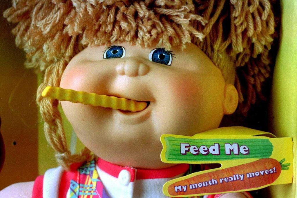 A doll with curly hair and blue eyes, holding a crinkle-cut fry in its moving mouth. It wears a colorful outfit. A label reads “Feed Me” with a carrot, and the text below says, “My mouth really moves!”.