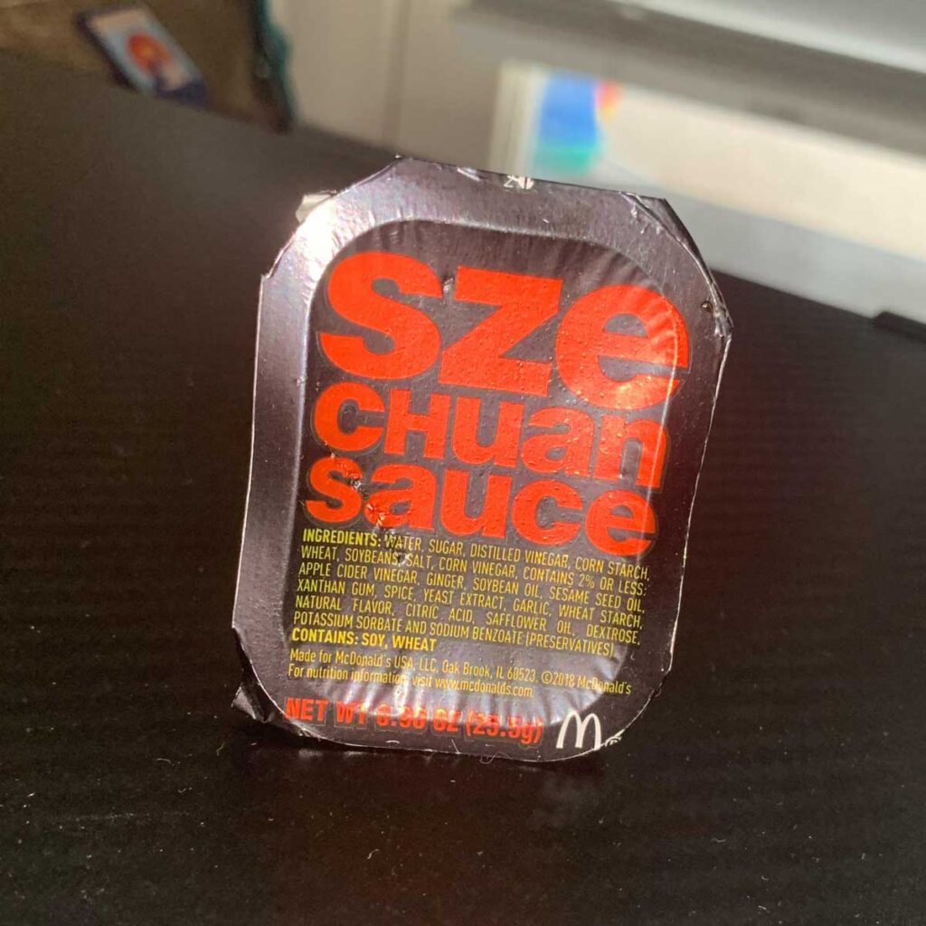 A McDonald's Szechuan sauce packet is displayed, featuring bold red text on a black background, reminiscent of discontinued fast food items that fans still crave. The ingredients and nutritional information are printed below the iconic sauce name.