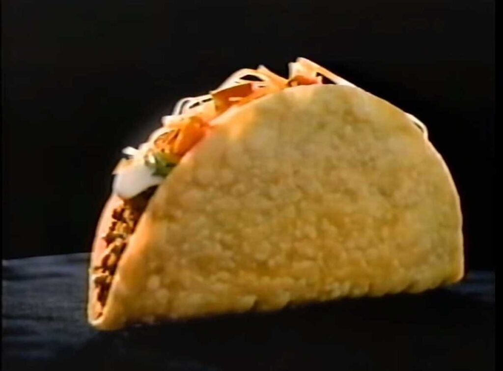 A crispy taco shell filled with seasoned ground meat, topped with shredded cheese, lettuce, diced tomatoes, and a dollop of sour cream—reminiscent of beloved discontinued Taco Bell menu items—set against a dark background.