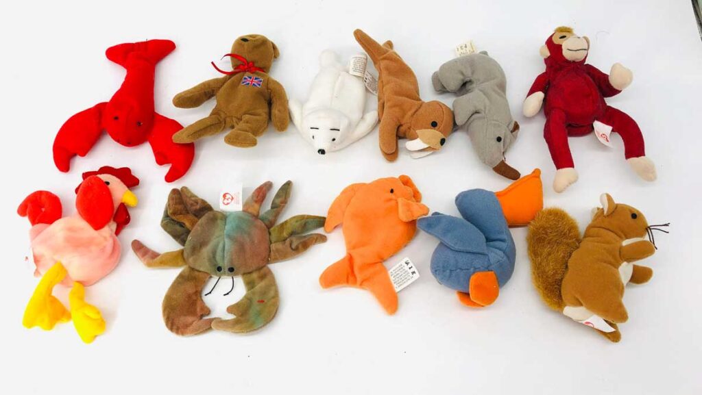 A collection of various plush toys, including animals like a lobster, bear, polar bear, monkey, rooster, crab, cow, seal, and squirrel, arranged on a white surface. Each toy is unique in color and design.