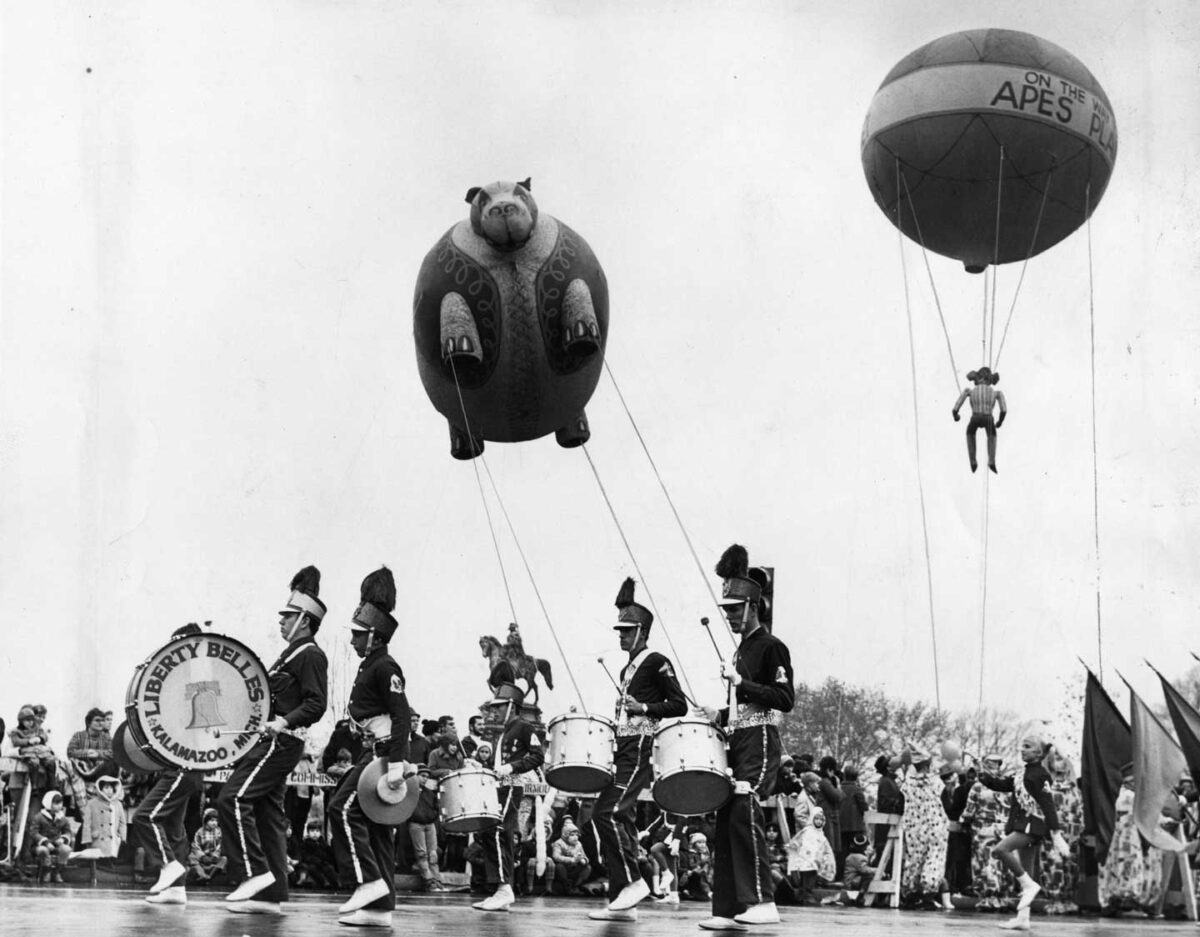 The nostalgic scene captures the Gimbel's Thanksgiving Day Parade, featuring a lively marching band with drummers in feathered hats. Above them, two whimsical balloons—a pig and an ape—float gracefully, enchanting a bustling crowd of spectators.