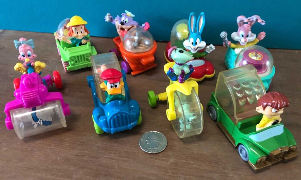 A collection of colorful toy cars featuring cartoon characters, displayed on a wooden surface with a quarter for scale. The toys vary in design and include characters seated in vibrant, imaginative vehicles.