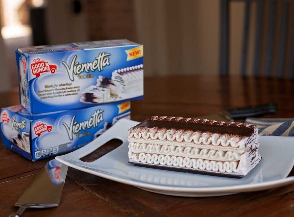 A Viennetta vanilla ice cream cake, a beloved treat reminiscent of discontinued snacks from the 90s, is elegantly arranged on a blue platter atop a wooden table. In the background, two unopened Viennetta boxes stand tall, with a serving knife resting beside the platter.