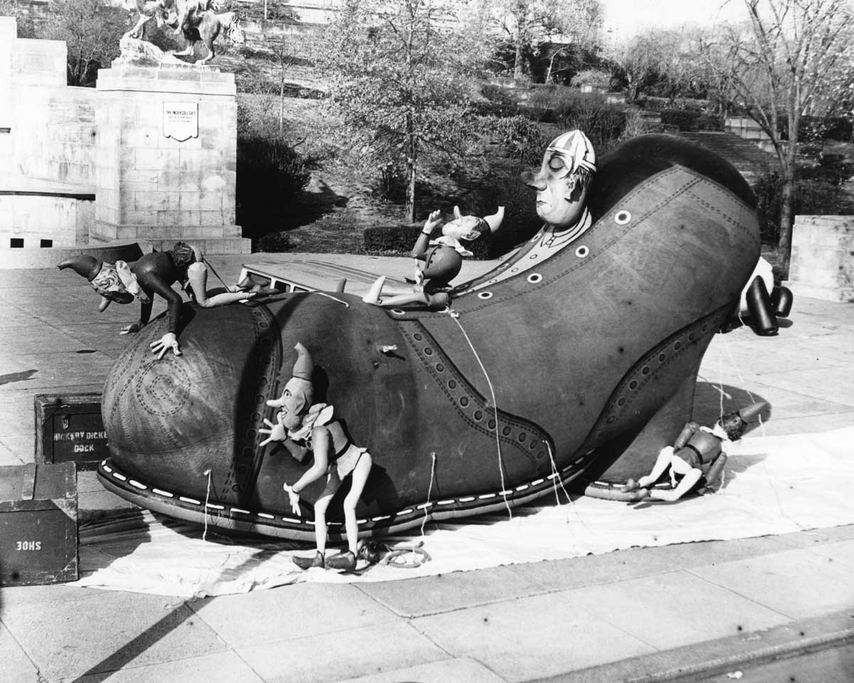 A giant shoe sculpture on a platform with whimsical figures climbing on it. The shoe features intricate details and a large face integrated into the design. Trees and stone structures are visible in the background.