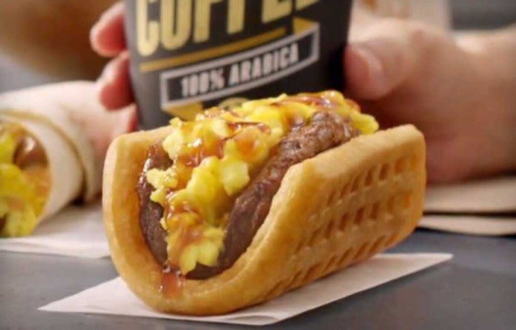 A breakfast taco, reminiscent of discontinued Taco Bell menu items, features scrambled eggs, sausage, and sauce in a waffle shell on a napkin. In the background, a hand gently cradles a steaming cup of coffee.