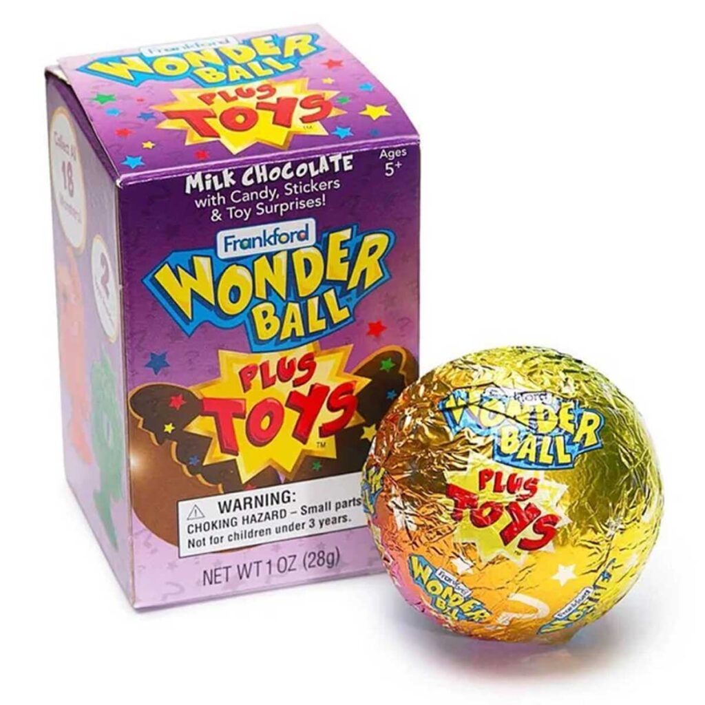 A Frankford Wonder Ball package sits beside an unwrapped Wonder Ball, reminiscent of discontinued snacks from the 90s. The colorful box features "Wonder Ball Plus Toys" text and a warning for small parts, while the chocolate treat inside gleams in its gold foil wrapping.