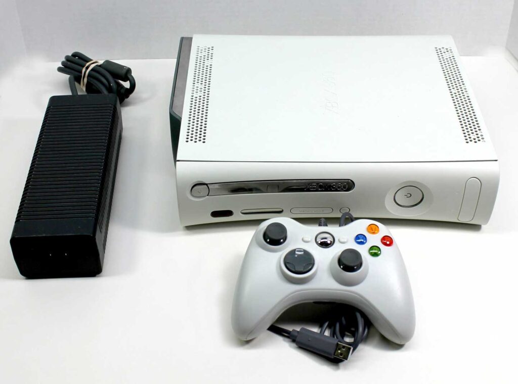 A white, best-selling video game console with a controller and power brick. The console includes vent holes on top and front, plus a disc slot, USB ports, and buttons. The controller sports multiple colored buttons, and the power brick has an attached cable.