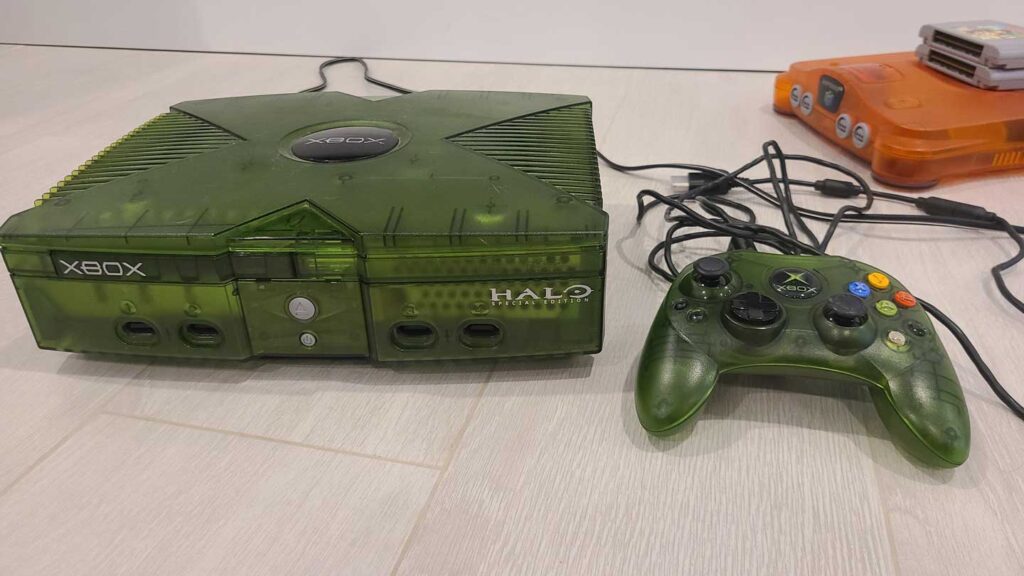 A translucent green Xbox console with a matching controller sits on a light wooden floor. With Halo branding, this console joins the ranks of best-selling video game consoles. In the background, an orange Nintendo 64 is partially visible.