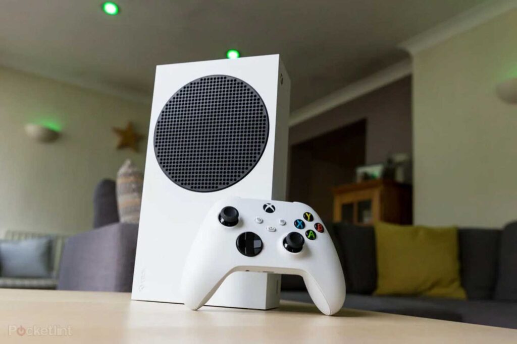 A white gaming console, one of the best-selling video game consoles, features a circular black vent and sits elegantly on a table beside a matching white controller. In the background, a cozy living room with couches and green accent lighting creates an inviting atmosphere.