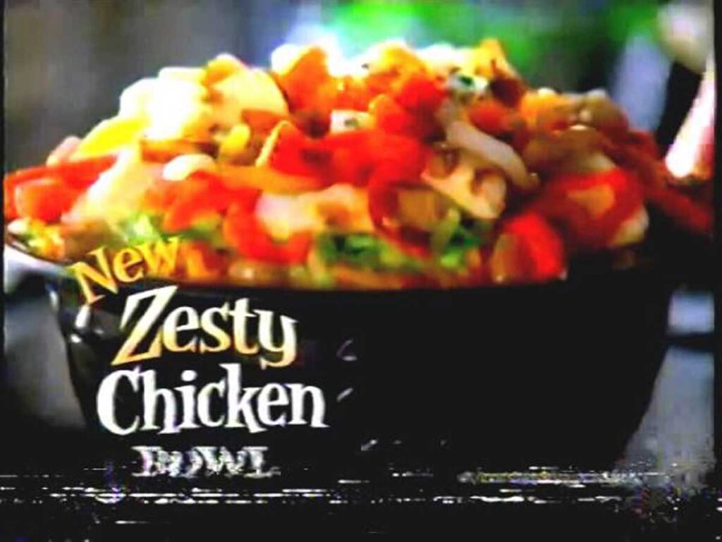 A colorful bowl of zesty chicken, reminiscent of discontinued Taco Bell menu items, is topped with diced tomatoes, cheese, and other garnishes. The text "New Zesty Chicken Bowl" is prominently displayed on the side of the bowl.