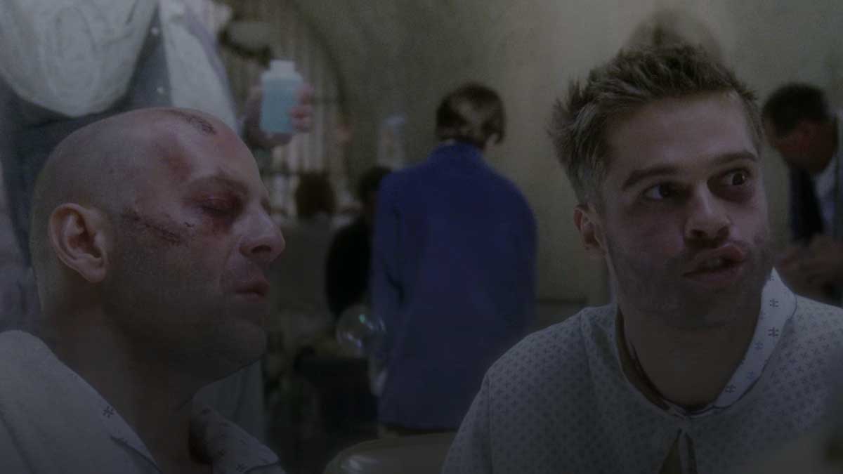 Two men in hospital gowns sit together in a dimly lit room. One man has a shaved head and facial bruises. The other has spiky hair and is speaking animatedly. People are blurred in the background, adding to the tense atmosphere.