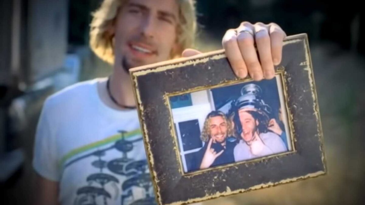 Songs turning 20 in 2025 - Nickleback Look At This Photograph. A person holds a framed photo featuring two people smiling and posing cheekily. One person in the photo wears a beverage container on their head. The holder wears a white graphic t-shirt and several rings. The scene is outdoors in daylight.