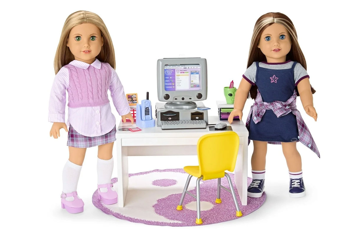 Two dolls stand beside a white desk with a retro computer, flip phone, and school supplies on top. The doll on the left wears a purple sweater, plaid skirt, and white socks. The one on the right wears a denim dress with a plaid shirt tied around the waist.